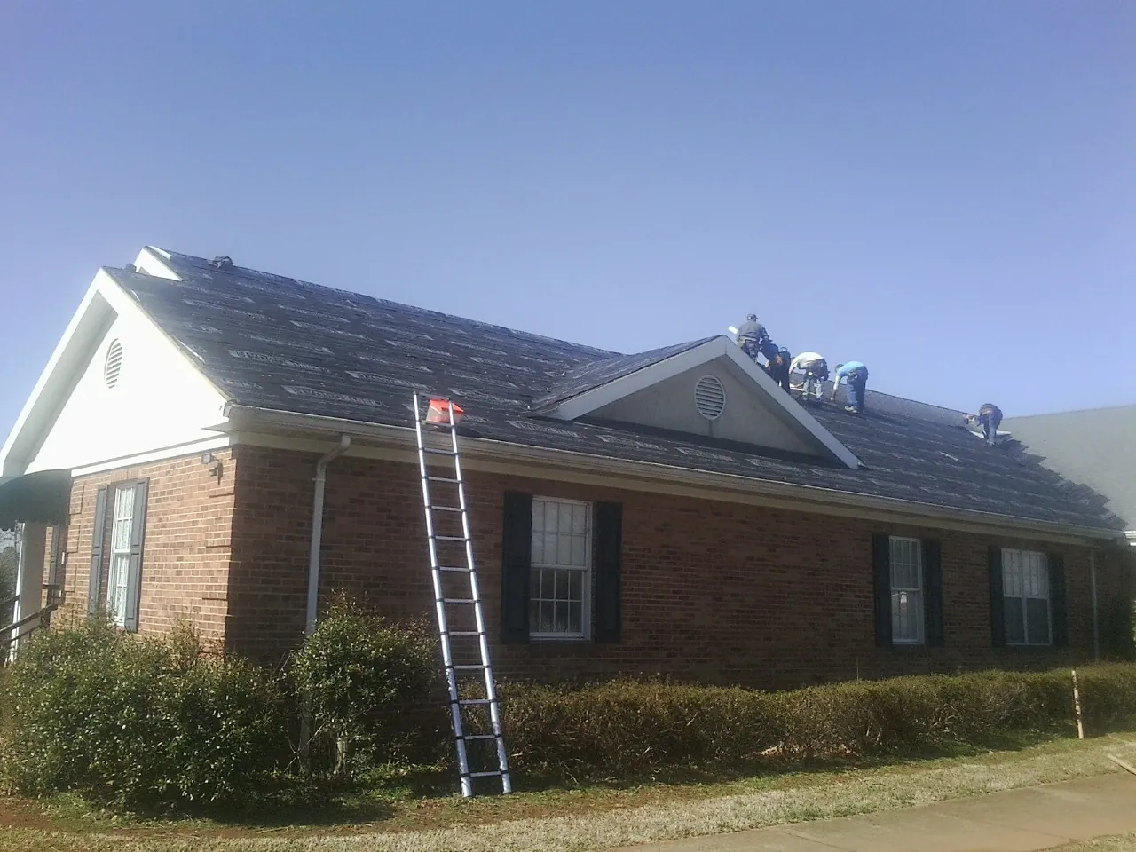 Crew tearing shingles off a brick ranch roof with a ladder set — Metal Roof Repair work by our crew in Neshkoro