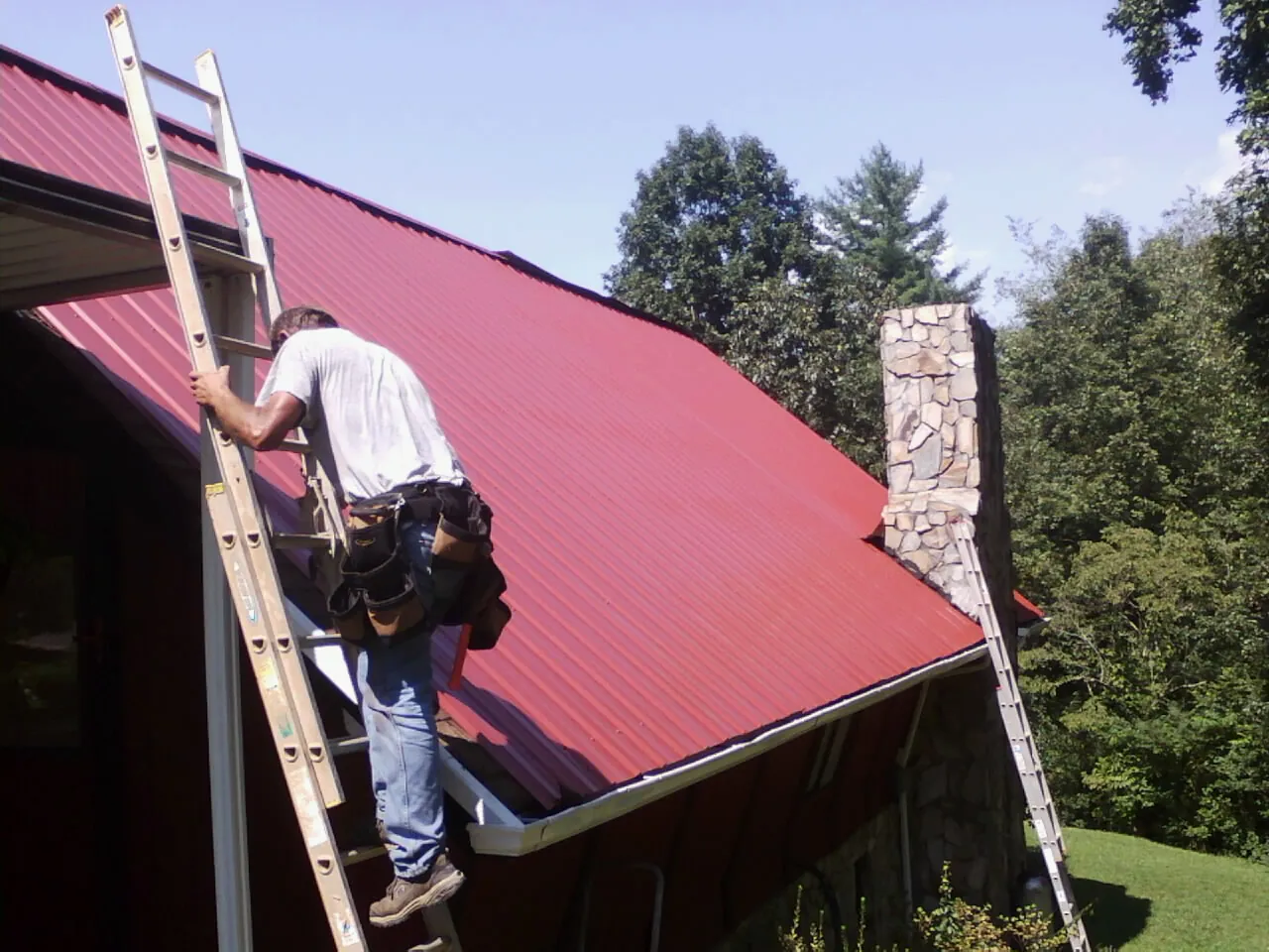 Roofer on a ladder beside a red metal roof and stone chimney — Soffit & Fascia Repair by Roofing Brothers in Neshkoro
